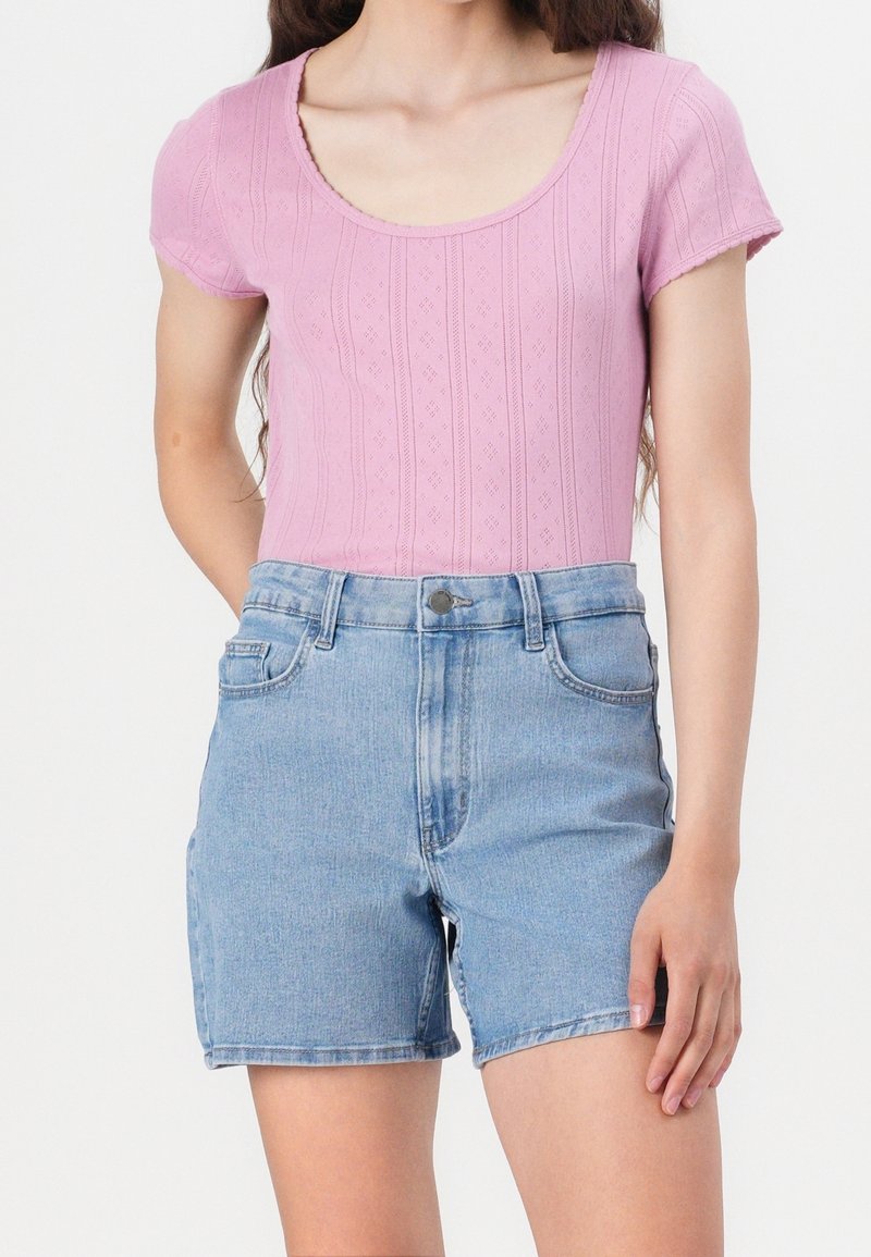 Femme portant un haut en maille côtelée rose à manches courtes, associé à un short en jean taille haute bleu clair, sur fond blanc.