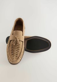 Beige mokasiner i suède med fläta på ovandelen, platt sula med blå accent och en dekorativ tofs framtill.