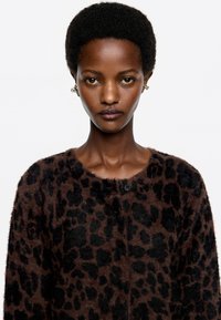 Cárdigan de estampado de leopardo marrón hecho de un tejido suave y texturizado con acentos negros, que cuenta con un escote redondo y cierre de botones.