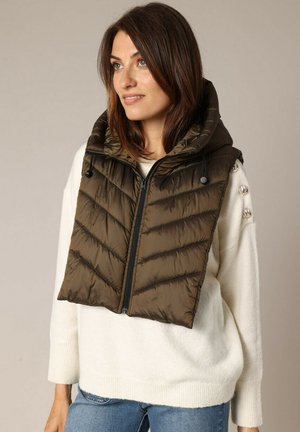 Femme aux cheveux bruns mi-longs portant une écharpe matelassée à capuche vert olive sur un pull blanc et un jean bleu, regardant vers la droite.