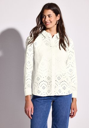 Camisa blanca de botones con intrincados patrones cortados, mangas largas y dobladillo curvado. Combinada con unos vaqueros de denim azul.