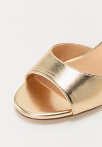 Gouden metallic slip-on sandaal met brede band, open teen ontwerp, zachte textuur en een platte zool. Heeft een zachte nude binnenvoering.