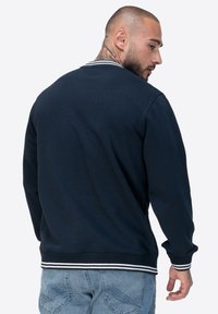 Marine Sweatshirt mit geripptem Kragen und Bündchen, versehen mit weißen Streifen. Der Stoff erscheint weich und hat eine lockere Passform, ergänzt durch hell gewaschene Jeans.
