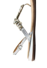 Sandalo piatto argento con decorazioni gioiello, cinturini testurizzati e un tacco basso e liscio. Chiusura con fibbia e soletta di un leggero color tan.