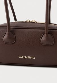 Braon kožni ručni torba s teksturiranim završetkom, zlatnim patentnim zatvaračem i dvostrukim drškama. Sadrži istaknuti "VALENTINO" logotip u zlatnoj boji.