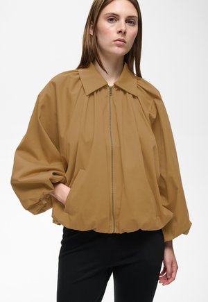 Femme portant une veste ample couleur sable à fermeture éclair, avec des manches bouffantes et un col, debout avec une main dans une poche sur un fond blanc.