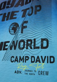 Blue cotton T-shirt featuring bold black text "THE TOP OF THE WORLD" and "CAMP DAVID." Includes stylized globe and wave pattern details.