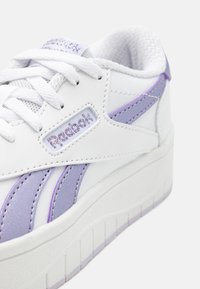 Bela usnjena superga s vijoličnimi poudarki in teksturirano površino. Ima dvignjen podplat, mrežasto podlogo in vezeno logotip na strani.