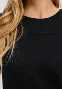 Musta pyöreäkaulatoppi, joka on valmistettu sileästä, joustavasta kankaasta. Sisältää huomaamatonta ommeltua yksityiskohtaa rinnassa. Yksinkertainen muotoilu ilman kuvioita.