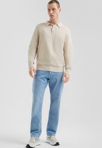 Beige strukturierter Langarm-Polo mit drei Knöpfen, kombiniert mit hellblauen geraden Jeans und weißen Sneakern mit grauen Akzenten.