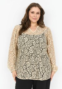 Kleitkanga pluus musta lillemustriga ja beež aluskihiga. Omab puffi varrukaid ja V-kujulist kaelust. kantud mustade pükstega.