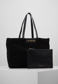 Svart tote-väska med fluffig textur och dubbla handtag, med en logoplatta. Följd av en slät svart påse med guldlock.