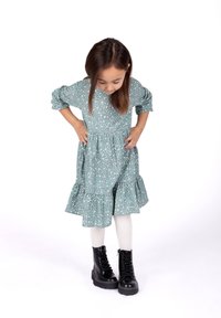 Ein türkisfarbenes Blumenkleid mit Puffärmeln und einem Rüschensaum, kombiniert mit schwarzen Schnürstiefeln und weißen Strumpfhosen.