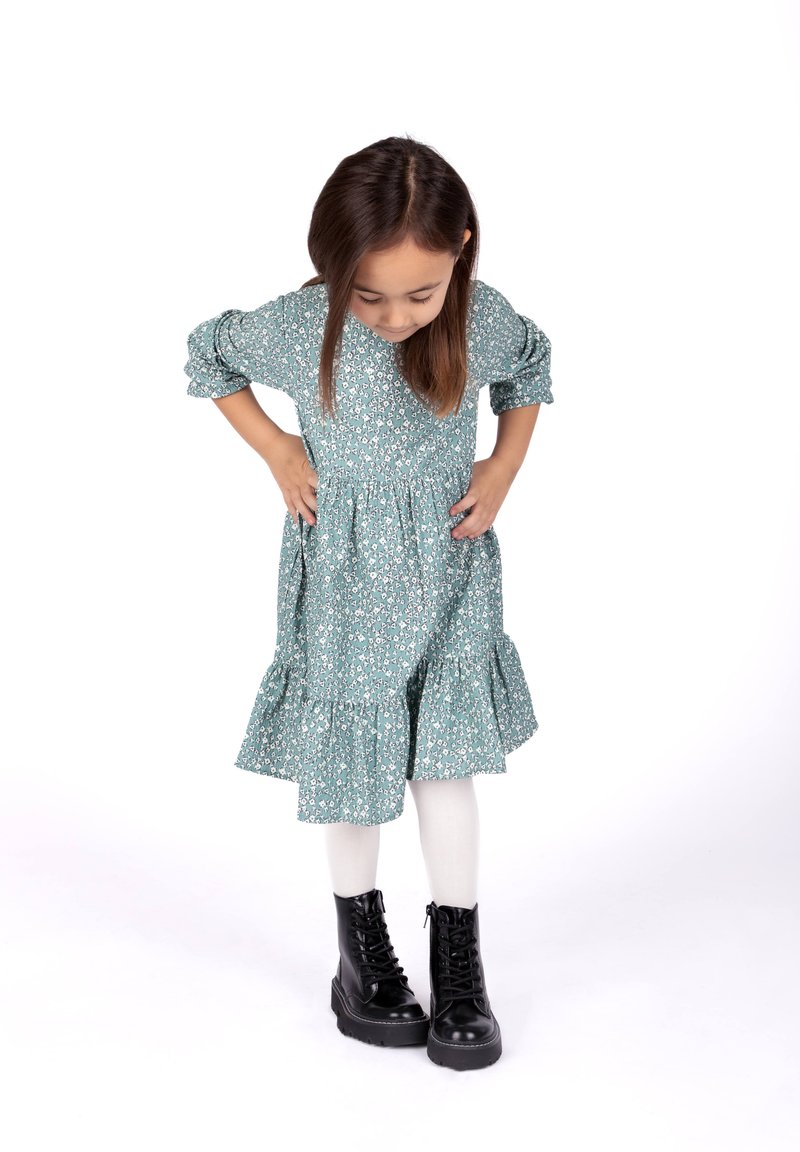 Ein türkisfarbenes Blumenkleid mit Puffärmeln und einem Rüschensaum, kombiniert mit schwarzen Schnürstiefeln und weißen Strumpfhosen.