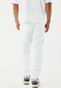 Persoon in witte sweatpants en een wit shirt, van achteren gezien, staand met voeten in zwart-witte sneakers op een witte achtergrond.