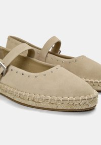 Beige Wildleder-ESPADRILLES mit flacher Sohle, rundem Zehenbereich, dekorativen silbernen Nieten entlang des Obermaterials und einem Riemen mit einer silbernen Schnalle.