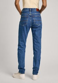 Μπλε τζιν παντελόνι σε στενή γρα�μμή, με σχέδιο πέντε τσεπών και δερμάτινη ετικέτα στη μέση στην απόχρωση του καφέ. Το ύφασμα έχει ελαφρώς ξεθωριασμένη υφή.