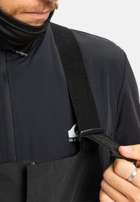 Close-up van een persoon die een zwarte verstelbare riem aanpast op zwarte buitenkleding met een rits en een klein logo op de borst.