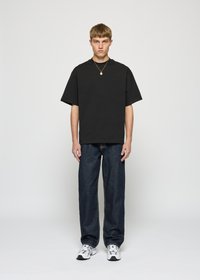 Young man standing wearing a black oversized T-shirt, dark blue jeans, white sneakers, and a gold necklace against a plain light background.