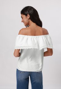 Weißes Off-Shoulder-Oberteil mit Spitzenbesatz, texturiertem Stoff und erhobenen Punkten, kombiniert mit blauen Jeans. Sichtbarer Rückenausschnitt und kurze Ärmel.