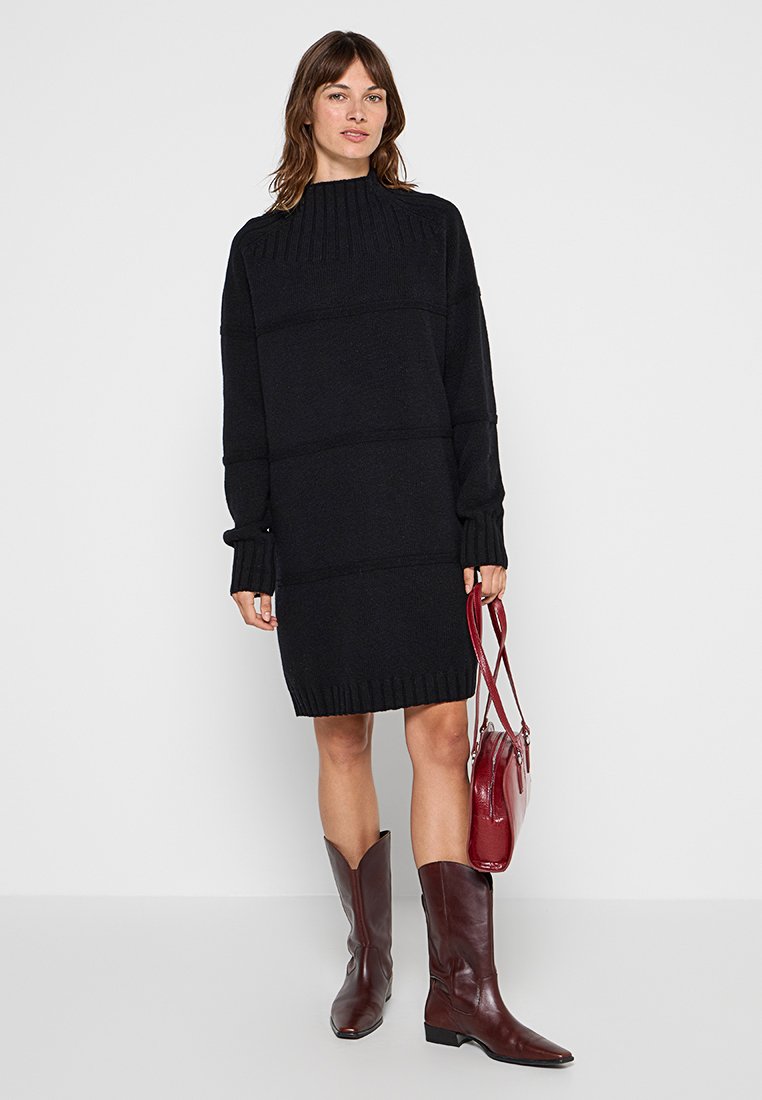 Femme portant une robe pull noire côtelée, tenant un sac à main rouge foncé, et chaussant des bottes hautes assorties rouge foncé.