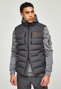 Grijze gewatteerde vest met een ritssluiting aan de voorkant, twee borstzakken en een capuchon. Voorzien van een logo patch en rode ritspulls. Gemaakt van een gestructureerde stof.