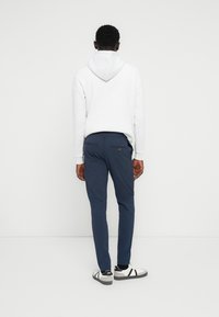 Άνδρας που φοράει λευκό φούτερ με κουκούλα, μπλε παντελόνι και λευκά αθλητικά παπούτσια, στέκεται απέναντι σε έναν λευκό τοίχο με τα χέρια χαλαρά στα πλάγια.