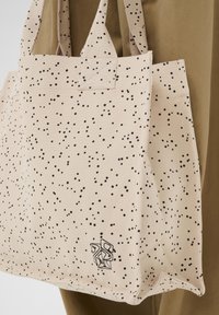 Sac fourre-tout en toile beige avec motif tacheté noir et logo abstrait, tenu par des poignées sur un fond en tissu marron.