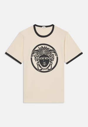 Chemise beige à manches courtes avec bordure noire au col et aux manches, présentant un grand motif noir de tête de Méduse centré sur le devant.