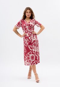 Rotes Blumenwickelkleid mit kurzen Ärmeln, das weiße Motive aufweist. Das Kleid hat eine gebundene Taille und einen knielangen Saum. Trägt beigen Absatzschuhe.