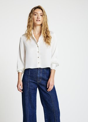 Blouse en satin blanc avec boutonnage sur le devant et manches longues, associée à un pantalon large en denim bleu foncé taille haute avec coutures contrastantes.