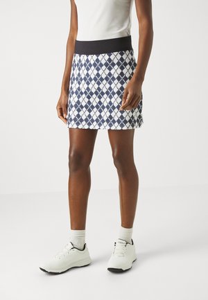 Persoon die een marineblauwe en witte argyle-rok draagt, een wit topje, witte sokken en witte sneakers, staand op een eenvoudige lichte achtergrond.