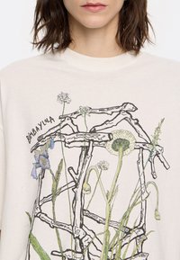 Cremfarbener T-Shirt mit einer botanischen Illustration von Blumen und Zweigen in verschiedenen Farben, mit "Bimbaylola" auffällig gedruckt.