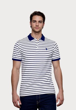 Bărbat tânăr purtând un tricou polo alb cu dungi orizontale bleumarin și guler bleumarin, asortat cu blugi bleumarin închis, stând în fața unui fundal simplu.