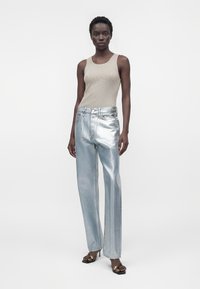 Mujer con top sin mangas beige, vaqueros plateados metálicos de talle alto y pierna ancha, y sandalias negras con tacón y detalle de cadena dorada.