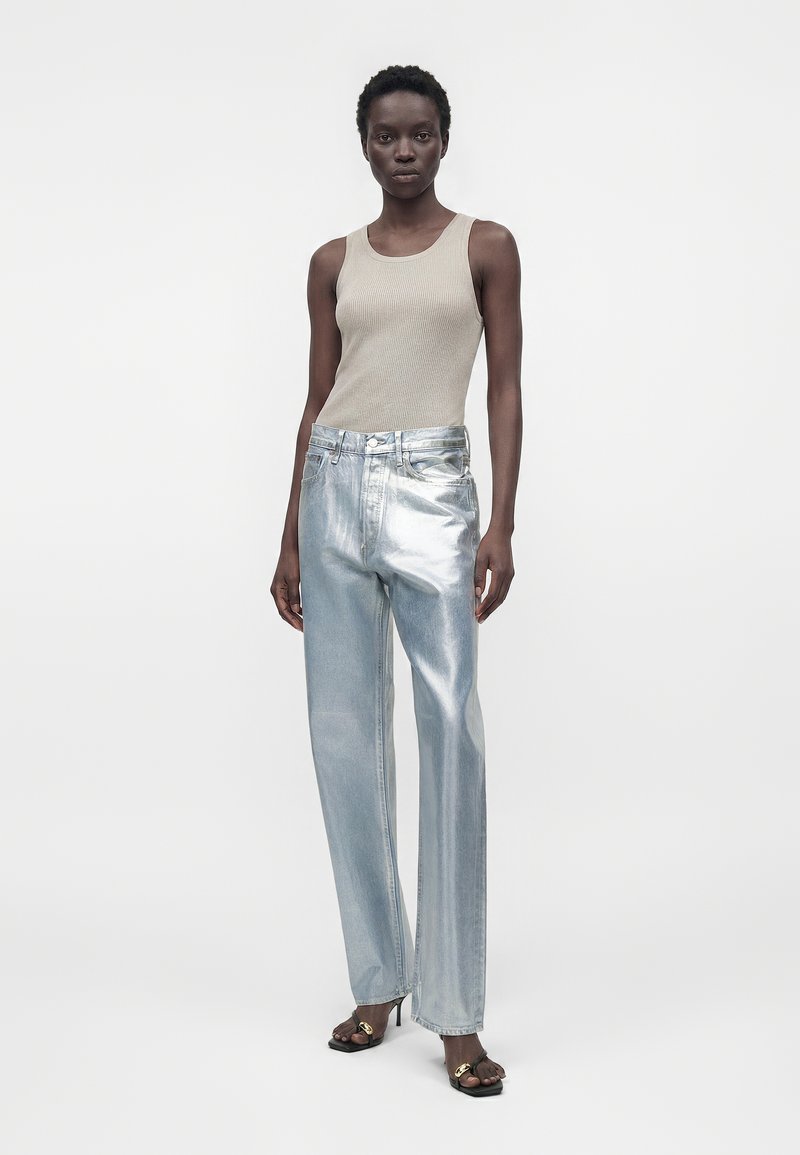 Femme portant un haut beige sans manches, un jean taille haute large en argent métallique et des sandales à talons noires avec un détail chaîne dorée.
