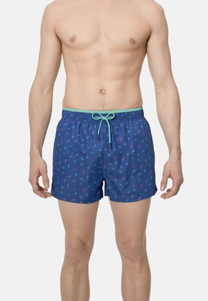 Hombre con pantalones cortos de baño azules con pequeños patrones de bicicletas coloridas y un cordón verde claro, de pie contra un fondo claro y liso.