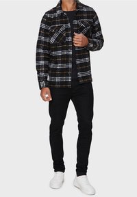 Chemise en flanelle à carreaux noir et blanc avec des accents orange, comprenant deux poches poitrine et une patte de boutonnage, portée avec un pantalon noir et des baskets blanches.