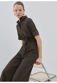 Frau in dunkelbraunem, mit Gürtel versehenem Jumpsuit mit hochgekrempelten Ärmeln, die seitlich auf einem transparenten Stuhl vor einem schlichten hellen Hintergrund sitzt.