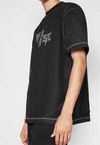 Svart mesh-t-shirt med rund hals, korta ärmar och kontrasterande vit sömnad. Grafisk design på framsidan ger visuell detalj.