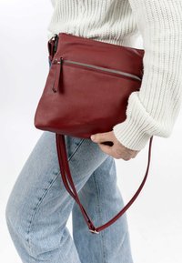 Rote Leder-Crossbody-Tasche mit einem Reißverschlussteil. Glatte Textur, rechteckige Form. Schlanker Schultergurt für einfaches Tragen befestigt.