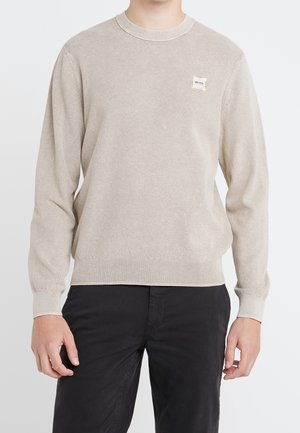 Mężczyzna ubrany w beżowy, teksturowany sweter z długim rękawem z małą naszywką z logo na klatce piersiowej oraz czarne spodnie, stojący na jednolitym tle.