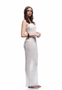 Vestido blanco, transparente y de punto con un patrón de diamantes y diseño sin tirantes. Accesorios con una pulsera oscura. Sandalias negras completan el conjunto.