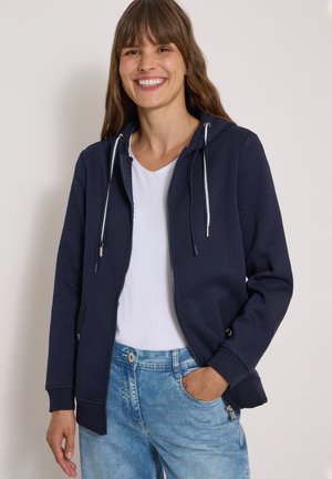 Femme souriante aux longs cheveux bruns portant un sweat à capuche bleu marine zippé, un t-shirt blanc et un jean bleu clair, une main dans la poche.