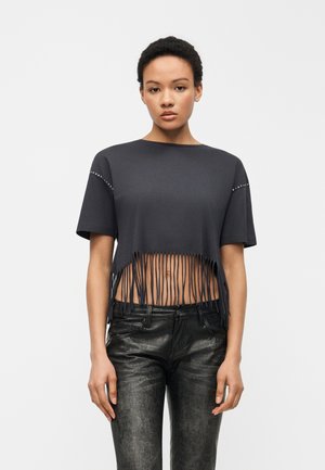 Jonge vrouw die een zwart cropped T-shirt draagt met franjes en studs op de mouwen, gecombineerd met zwarte, getextureerde leren broek.