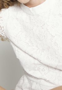 Haut en dentelle blanche avec col haut et motif floral, présentant des détails fins et un tissu texturé. Manches courtes, structure ajustée.