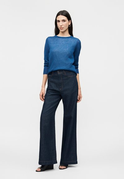 Femme aux cheveux longs et foncés portant un pull tricoté bleu, un jean large taille haute foncé, des chaussures noires à bouts ouverts et des boucles d'oreilles en forme d'anneaux dorés sur fond blanc.