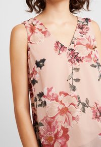 Vaaleanpunainen hihaton yläosa, jossa suuret punaiset ja tummanpunaiset kukat vihreillä lehdillä, V-pääntie, kevyt kangas.