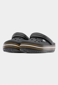 Ζευγάρι μαύρα παπούτσια τύπου clog χωρίς κορδόνια, με καφέ και λευκές ριγέ σόλες, οπές αερισμού και ρυθμιζόμενα λουράκια στη φτέρνα, εμφανιζόμενα πάνω σε γκρι φόντο.