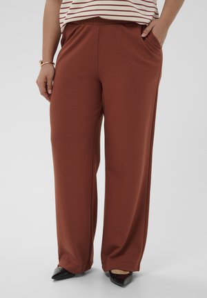 Persona che indossa pantaloni marroni a vita alta e a gamba larga, top a righe bianche e rosse, scarpe nere con punta appuntita, mani in tasca, su sfondo semplice.